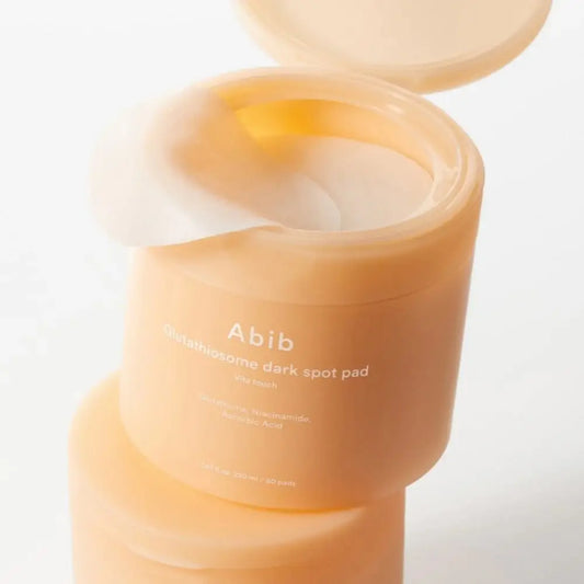 A peach-colored cylindrical container of abib glutathione dark spot pad holding translucent pads for brightening vitamin c toner use