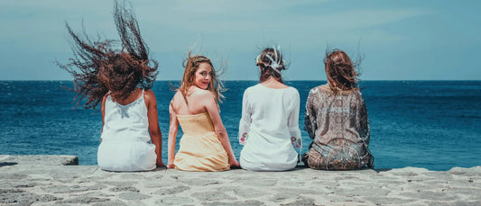 Four women in flowing light dresses sit back-to-back on a stone ledge overlooking the ocean for korean skincare south africa.